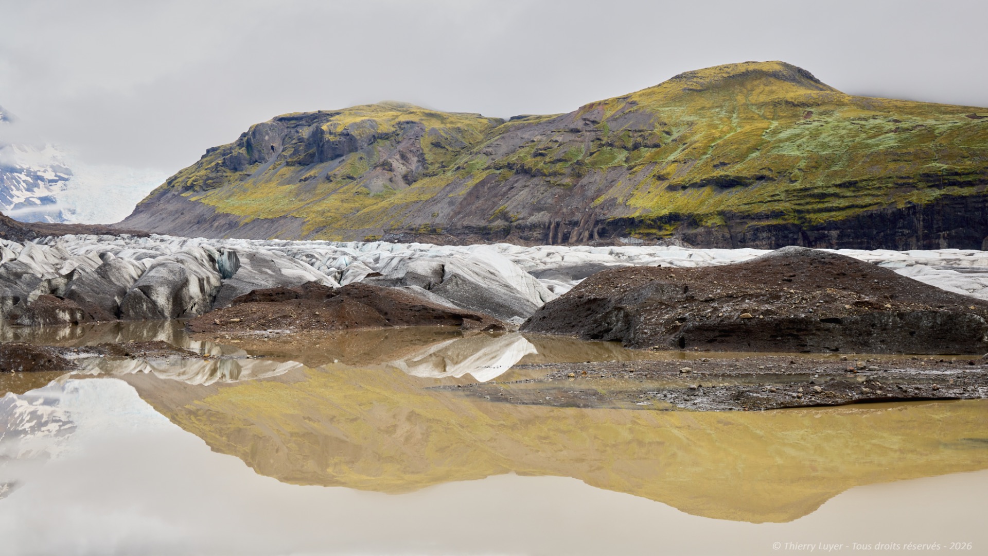 Glacier tongue and rocky mountains reflected in still water under an overcast sky.