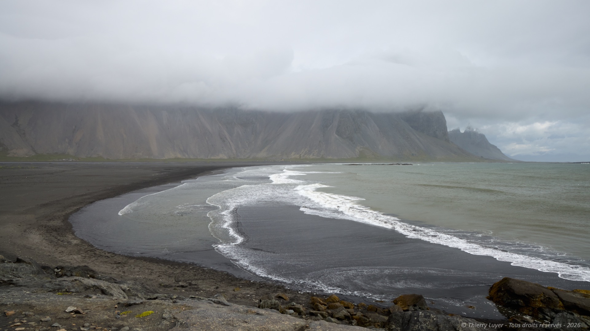 Moody coastline with black sand beach, foamy waves, and towering cliffs under overcast sky.