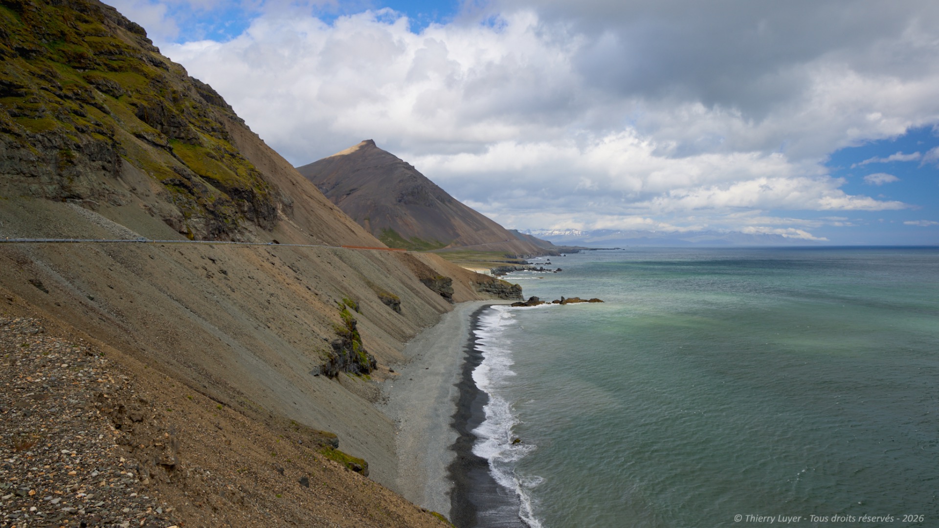 Coastal road curves along a black-sand beach beneath steep green mountains and cloudy blue sky.