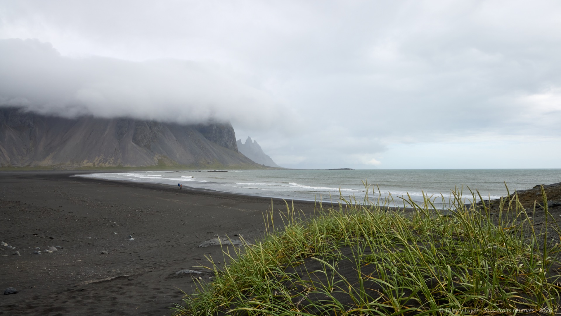 Black sand beach with grassy foreground, waves rolling in, and steep rocky mountains under cloudy sky.
