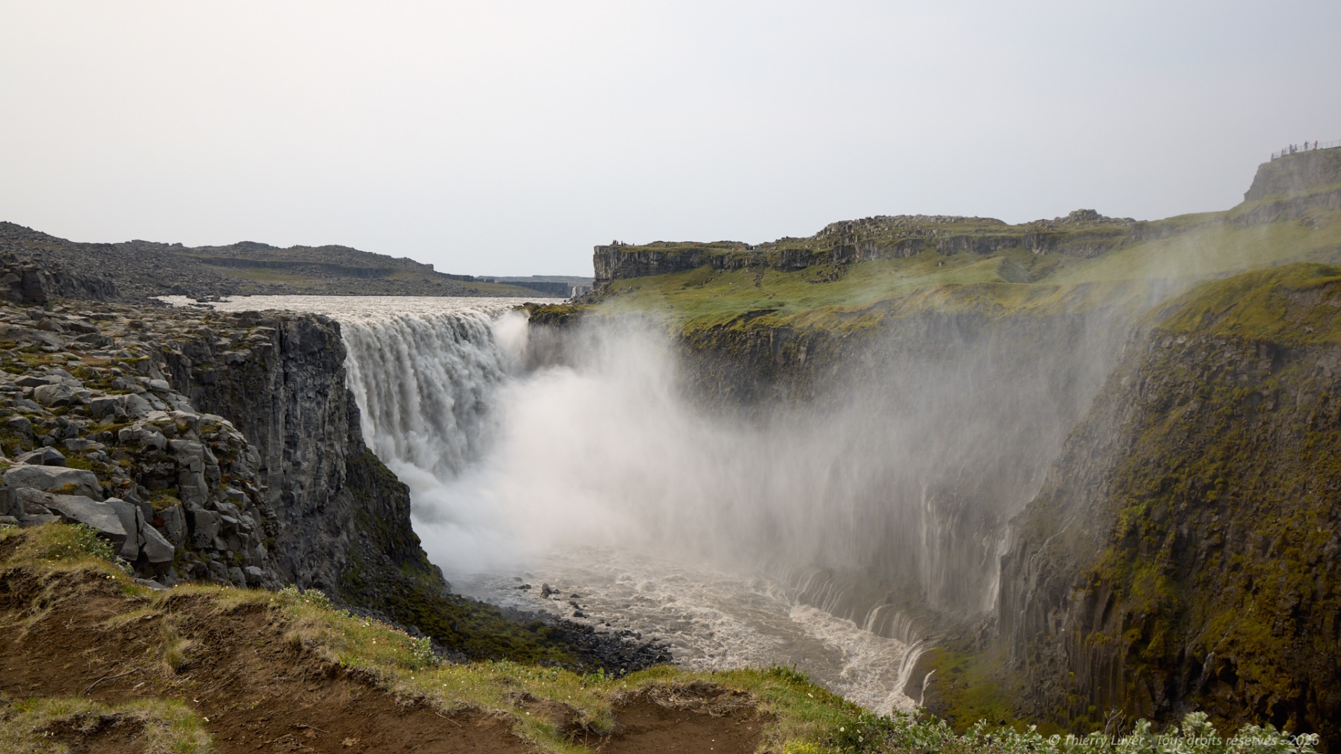 Powerful waterfall plunges over rocky cliffs, surrounded by green hills and misty spray under overcast sky.