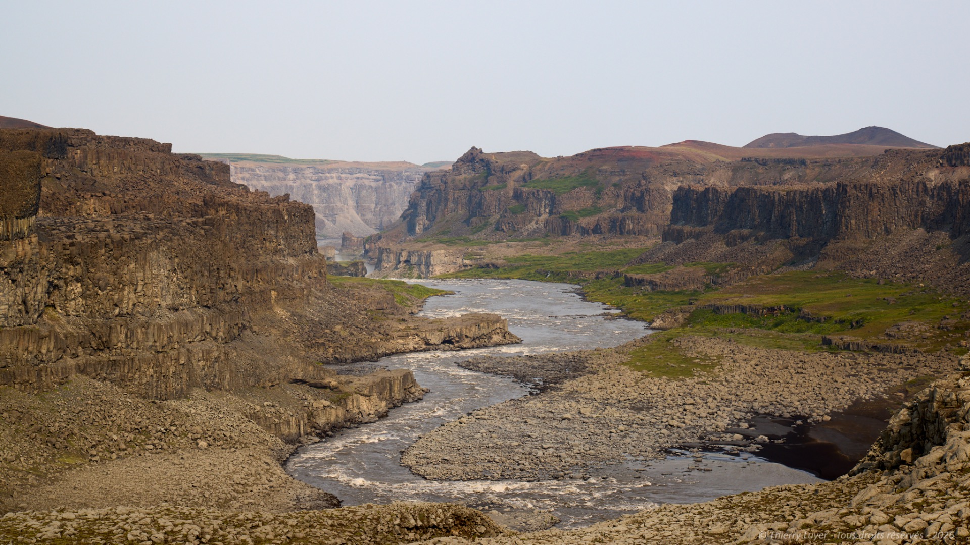 Rocky canyon landscape with a winding river, steep cliffs, and distant hills under a pale sky.