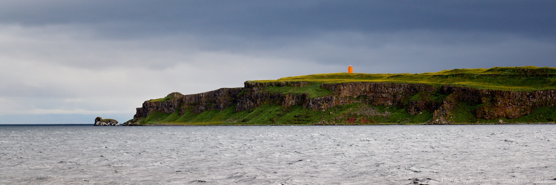 Cliffside lighthouse with orange top on grassy headland above choppy ocean under cloudy sky.