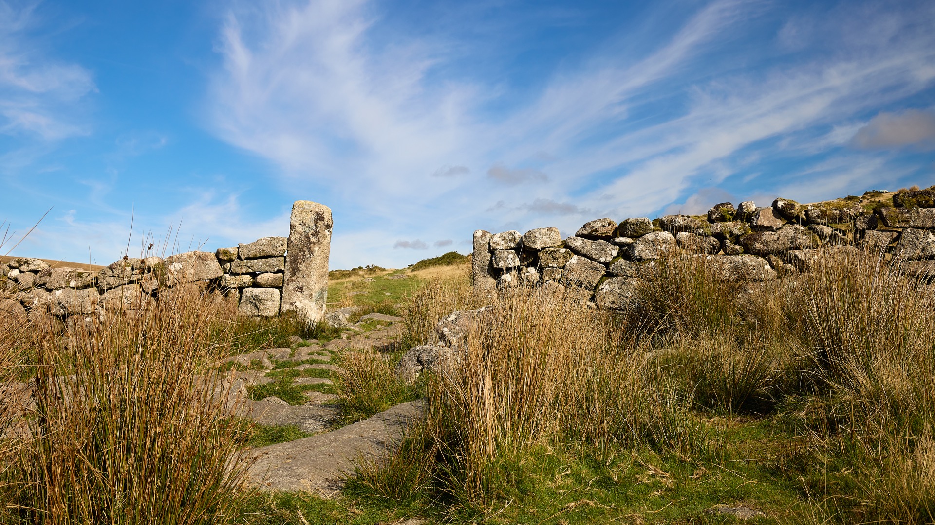 Sentier dans une lande verdoyante, pierres anciennes et murs de ruines sous un ciel bleu nuageux.