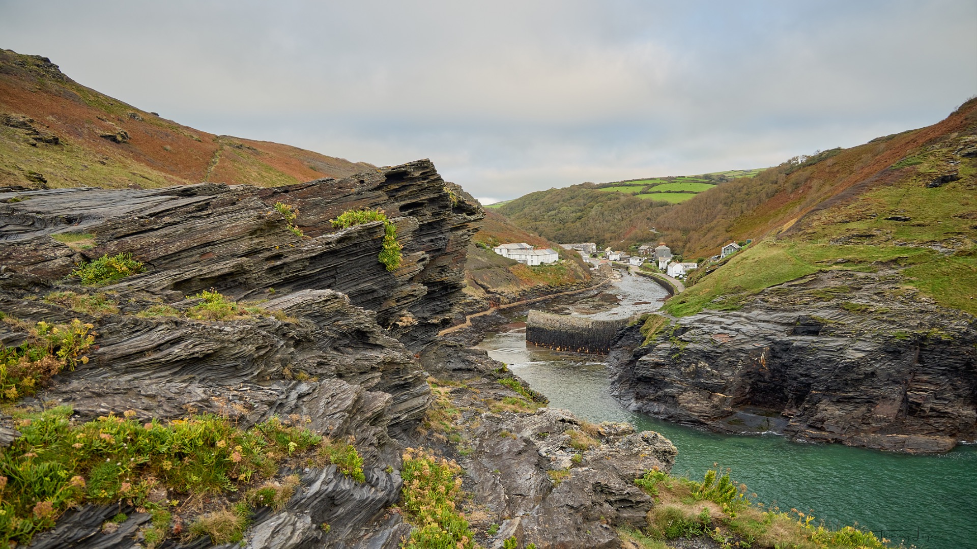 Falaises rocheuses stratifiées, rivière étroite et village de pêcheurs au fond, collines verdoyantes sous un ciel nuageux.