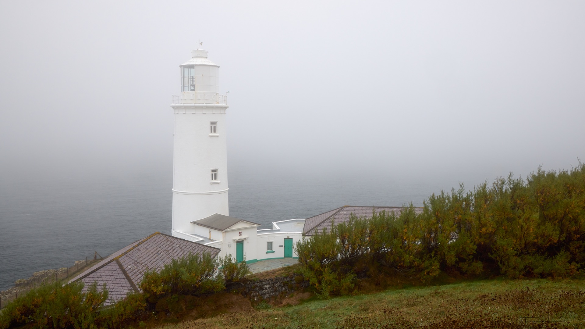 Phare blanc sur une falaise rocheuse, brume épaisse, vagues déferlant sur l’océan gris.
