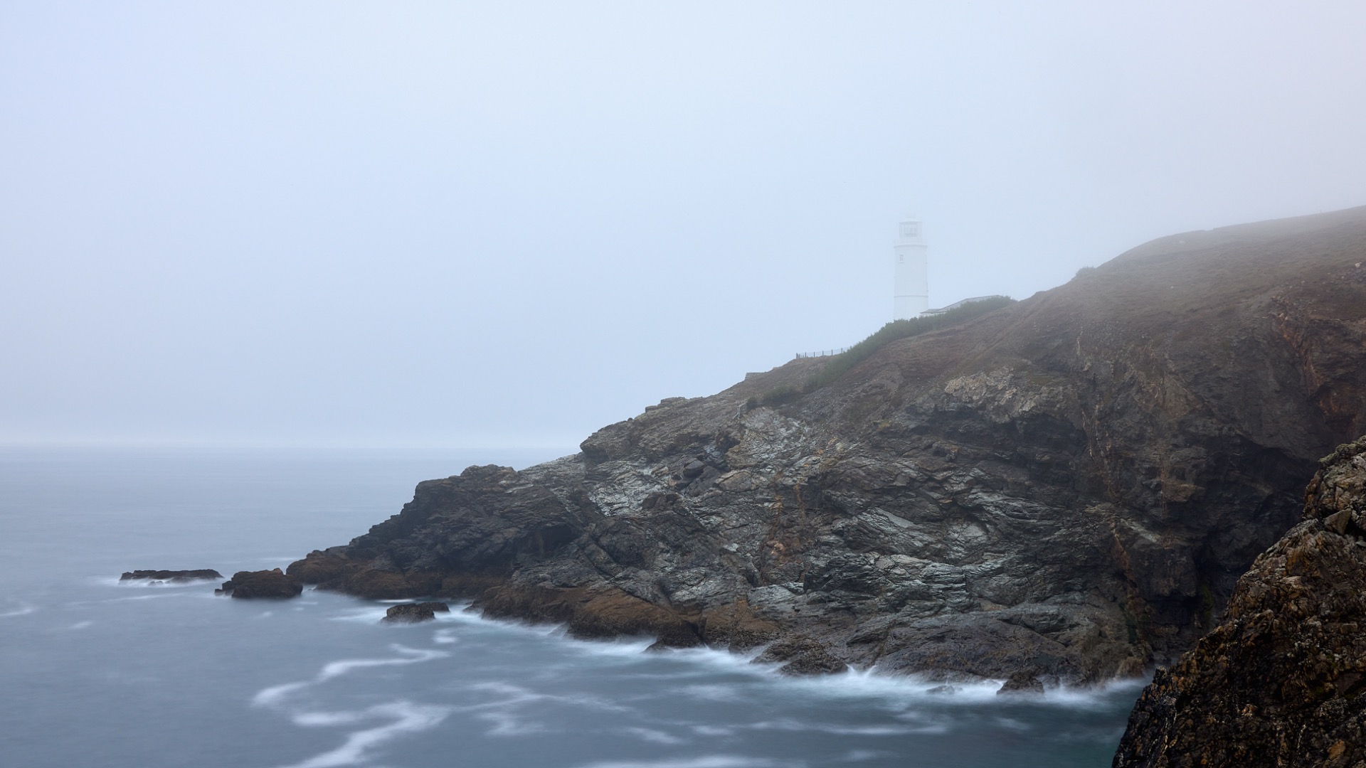 Phare blanc sur une falaise rocheuse, brume épaisse, vagues déferlant sur l’océan gris.