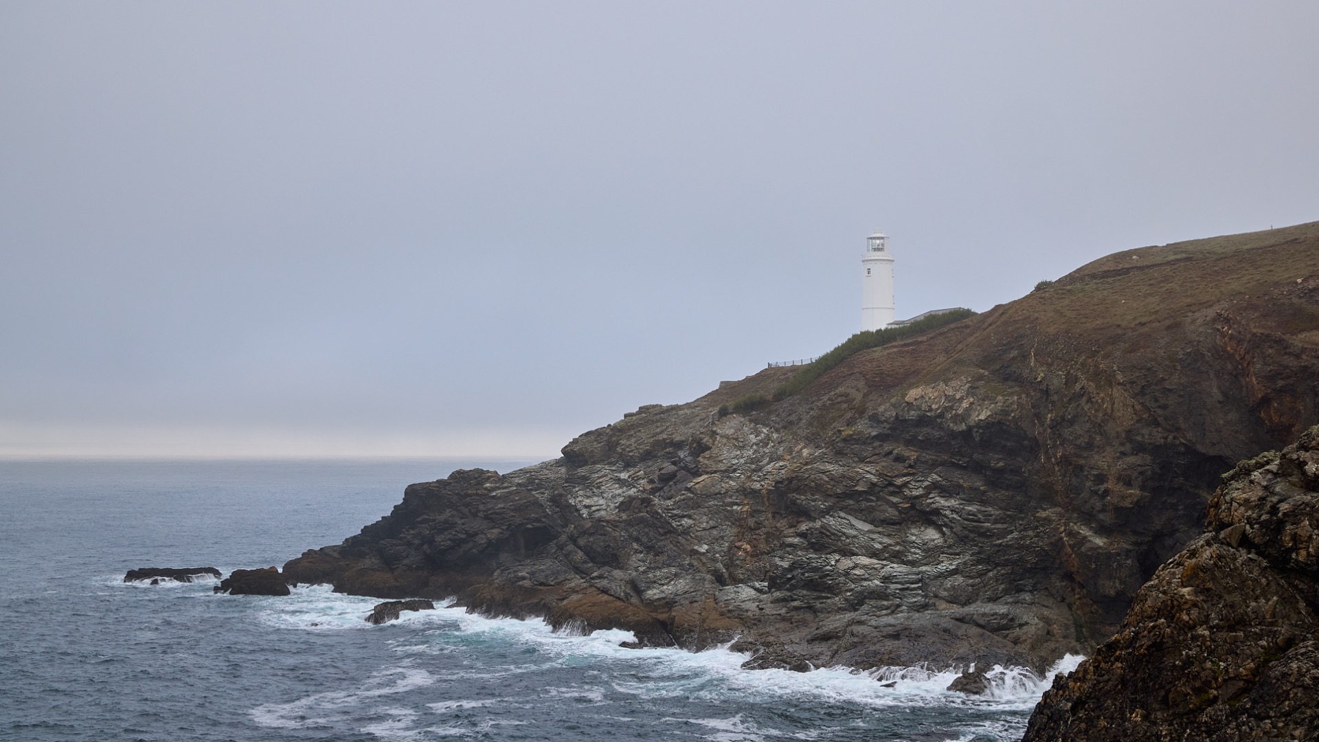 Phare blanc sur une falaise rocheuse, brume épaisse, vagues déferlant sur l’océan gris.