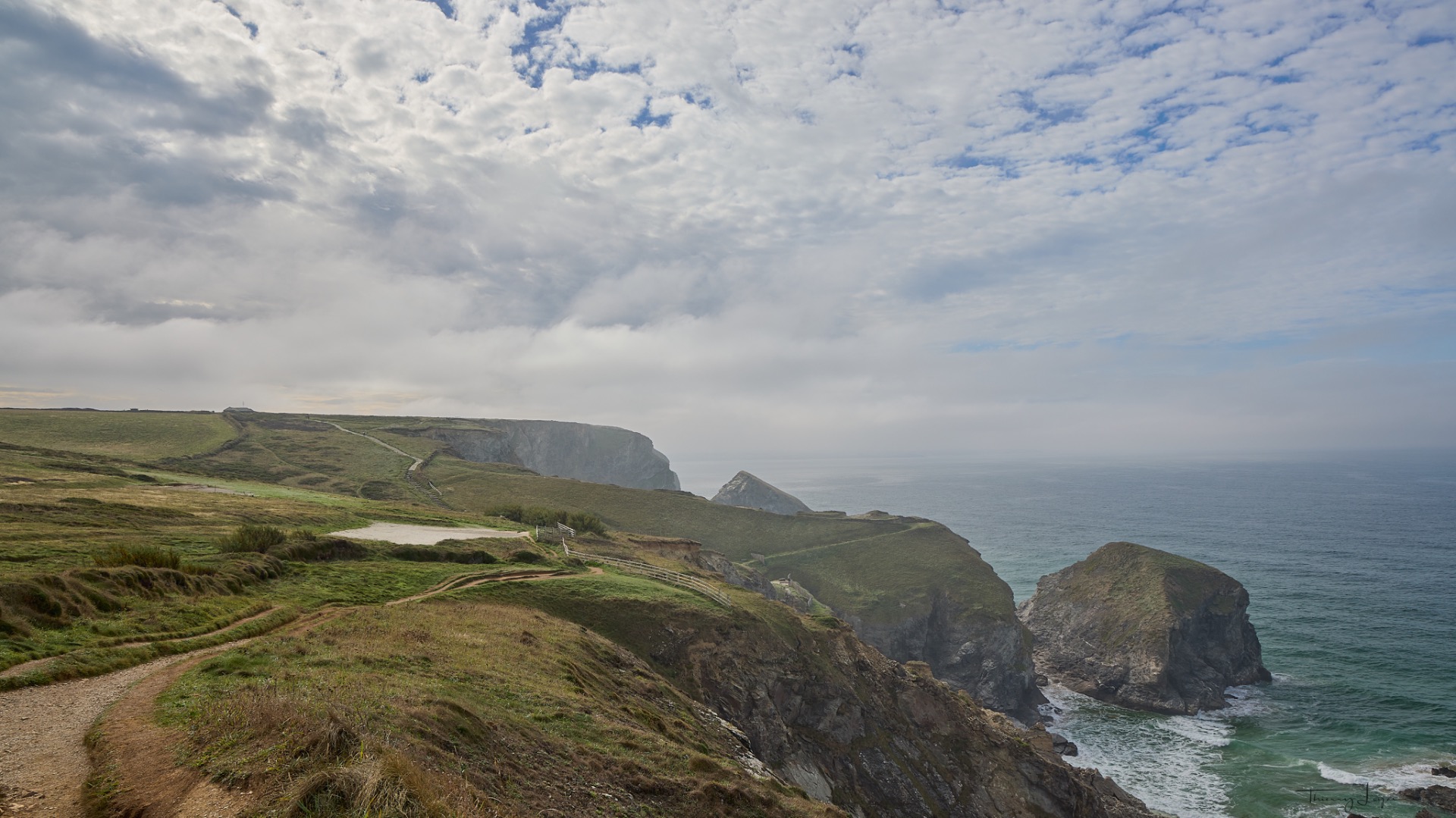 Falaises verdoyantes au bord de l’océan, sentier sinueux et clôtures, ciel nuageux dramatique.