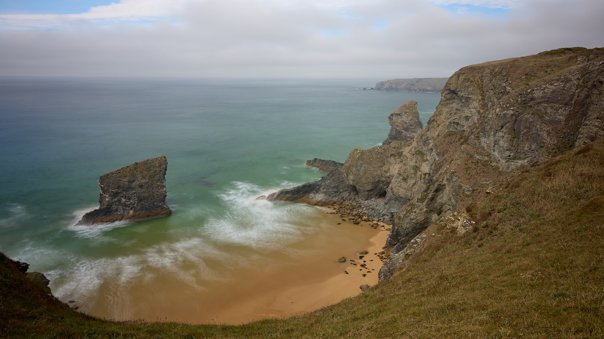 Falaises rocheuses surplombant une plage de sable, océan turquoise et vagues, ciel nuageux au loin.