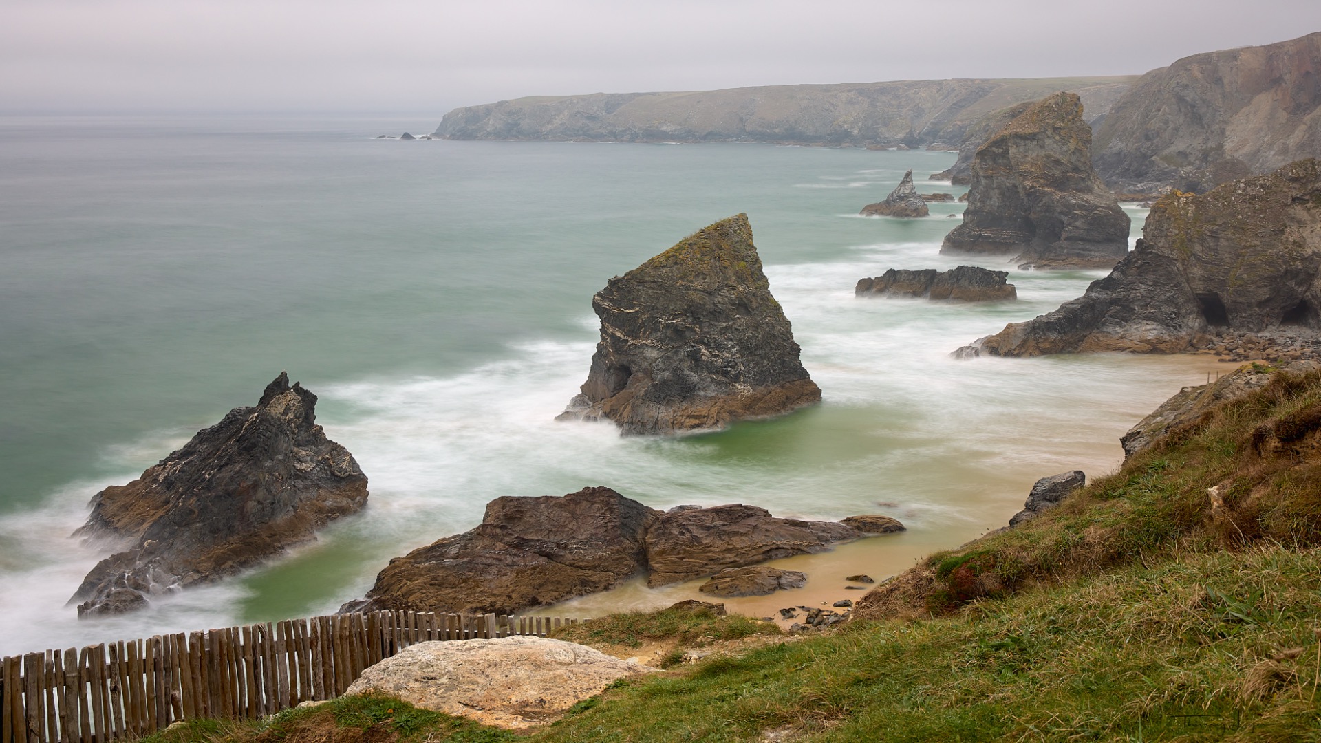 Falaises rocheuses et plage de sable, vagues déferlantes, clôture en bois au premier plan, mer brumeuse.
