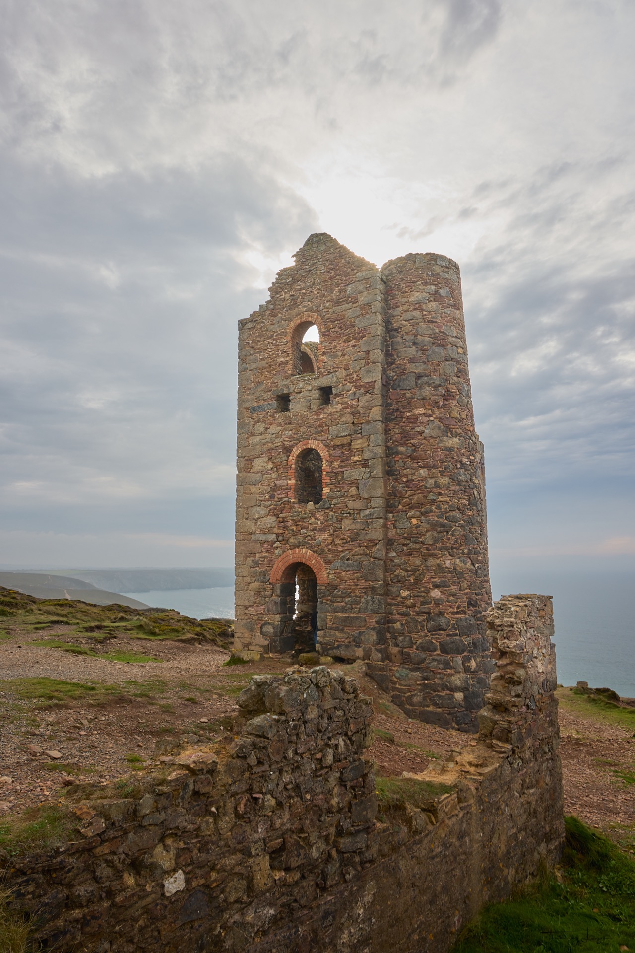 Tour en pierre en ruines avec arches et fenêtres, sur une côte verdoyante sous ciel nuageux.