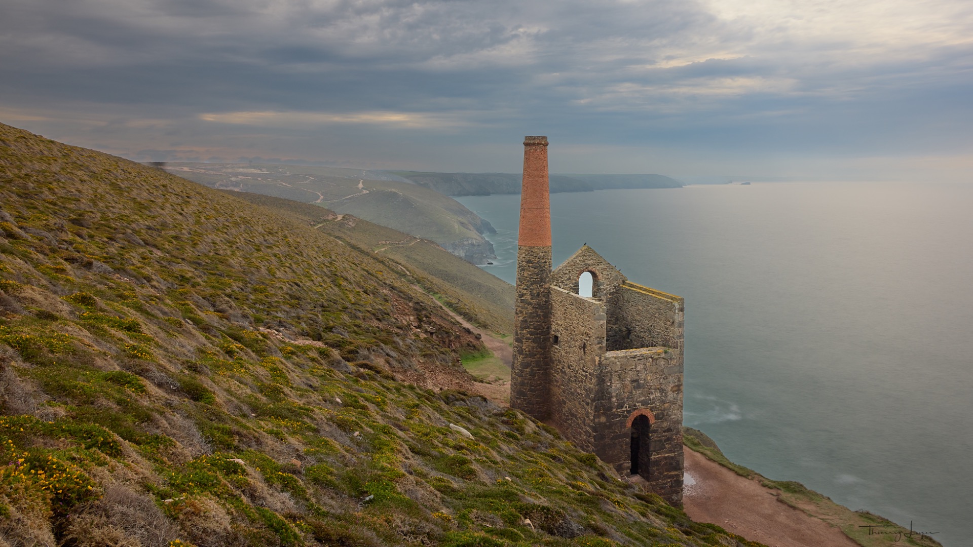 Mine d'étain en pierre en ruines, cheminée haute, sur une falaise côtière, mer calme et collines verdoyantes.