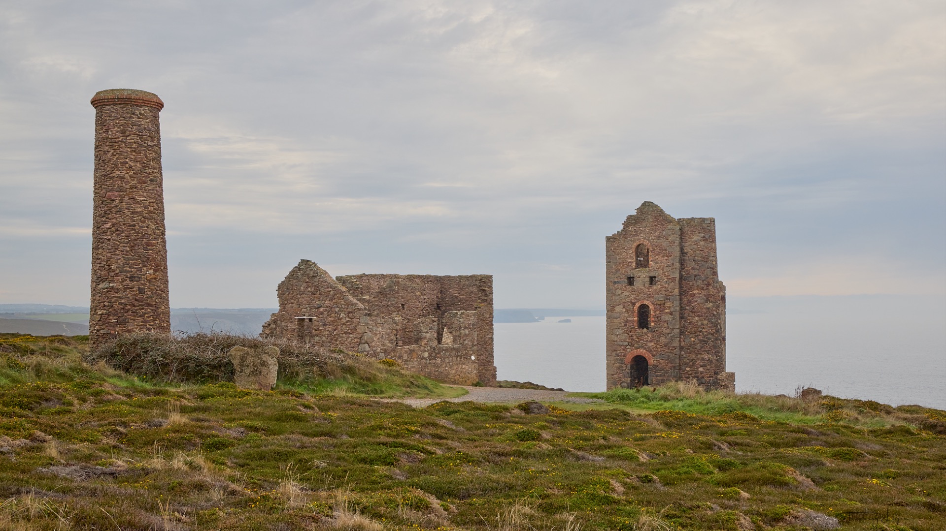Ruines de mines d'étain en pierre sur une falaise côtière, ciel nuageux et herbes vertes au premier plan.