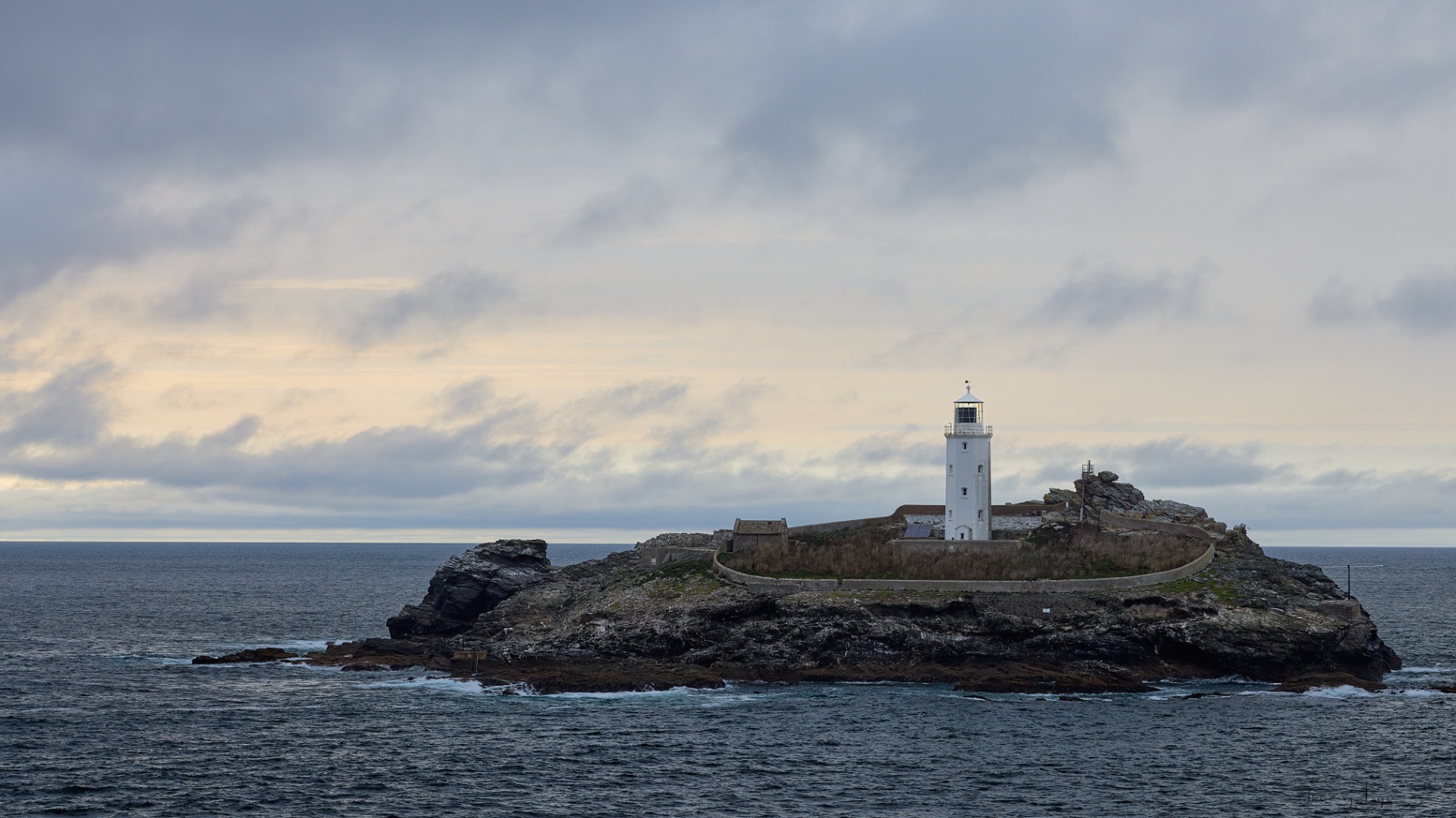 Phare blanc sur un rocher côtier, mer agitée et ciel nuageux au coucher du soleil.