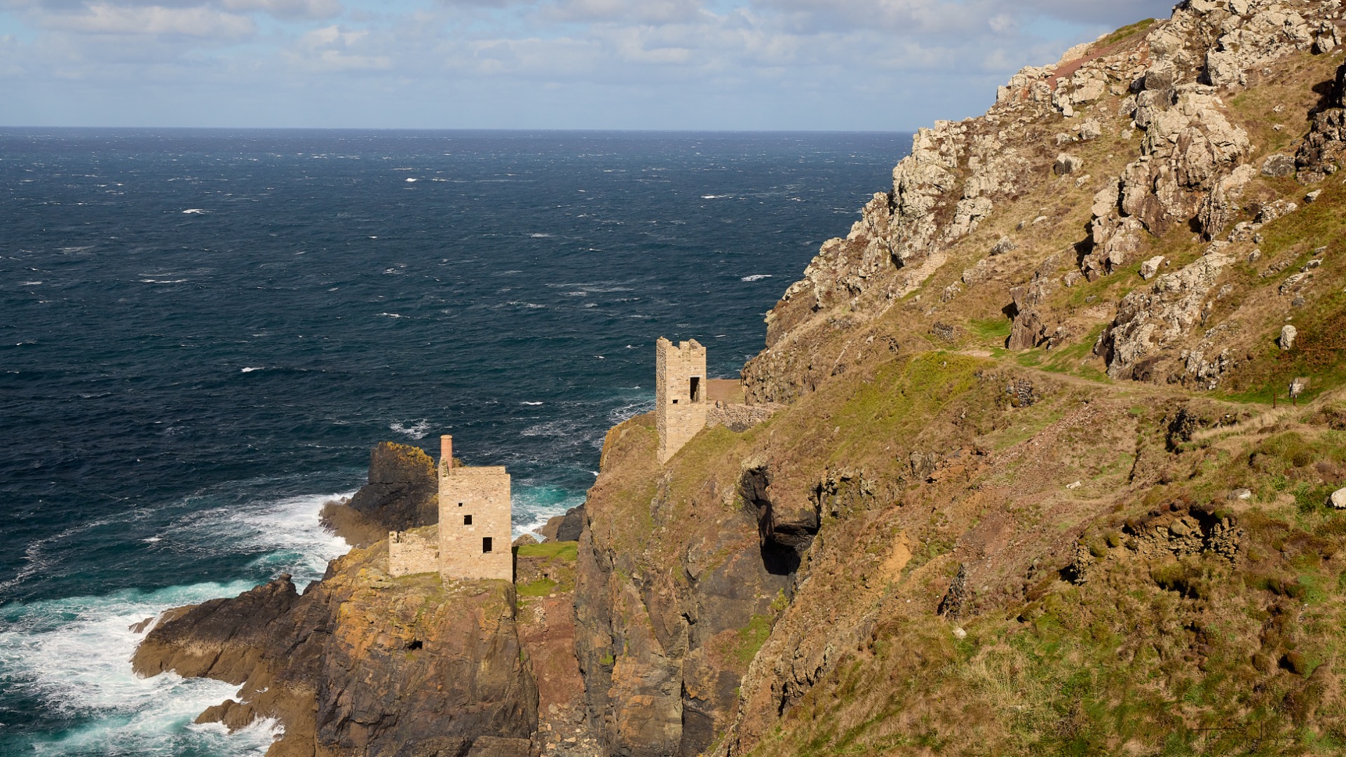 Falaises côtières verdoyantes avec deux tours en pierre ancienne dominant l’océan Atlantique, ciel nuageux.