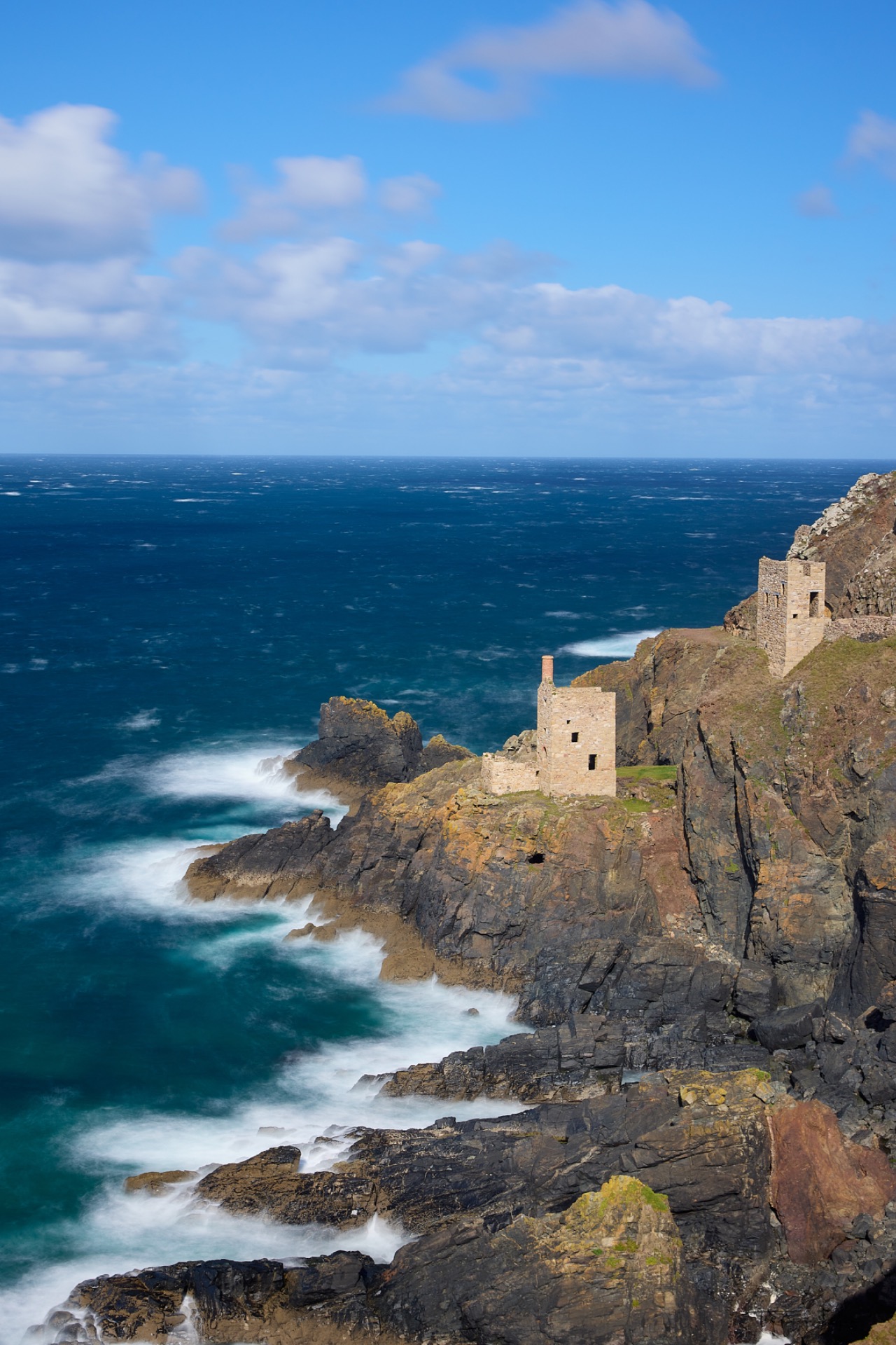 Falaises rocheuses au bord de l’océan, vagues écumantes et ruines de mines en pierre sous un ciel bleu nuageux.