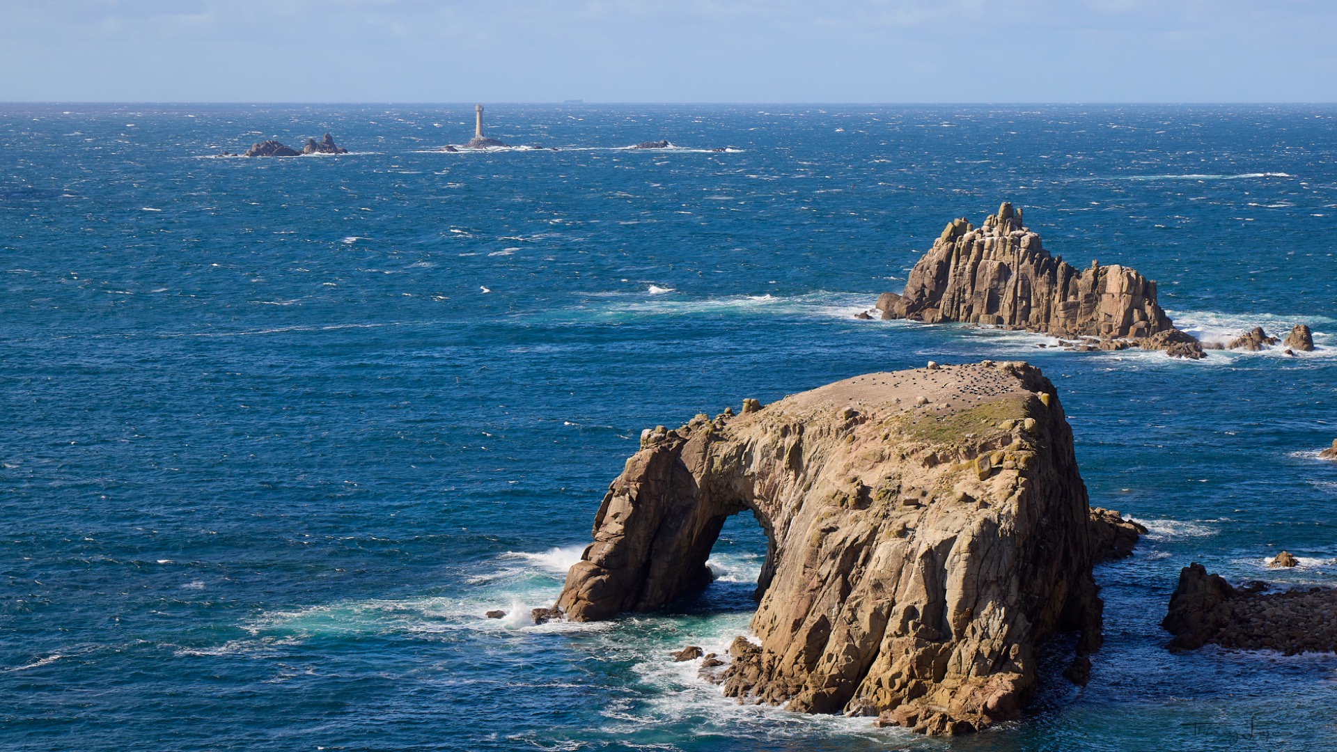 Océan bleu avec falaises rocheuses et arche, vagues écumantes et phare au loin sous un ciel clair.