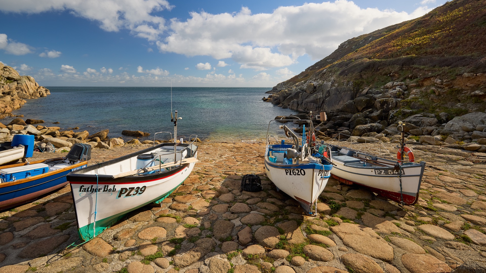 Trois petits bateaux de pêche amarrés sur des rochers, vue sur la mer et les falaises sous un ciel nuageux.