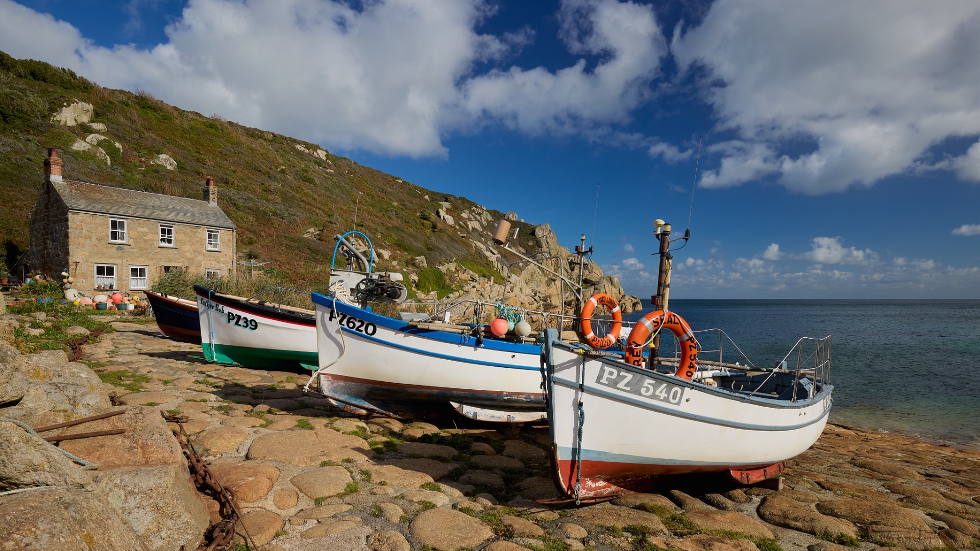 Bateaux de pêche amarrés devant une maison en pierre, côte rocheuse et ciel bleu nuageux.
