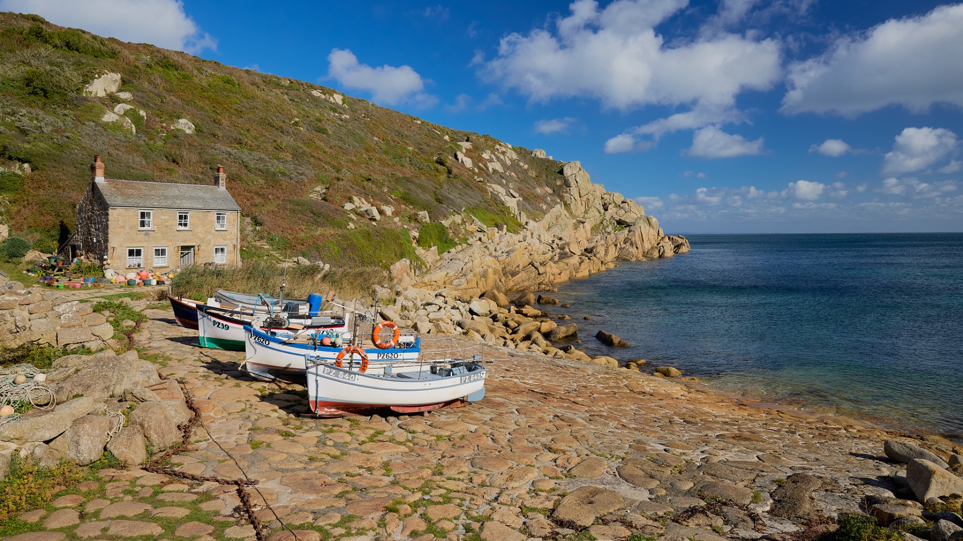 Bateaux de pêche amarrés devant une maison en pierre, côte rocheuse et ciel bleu nuageux.