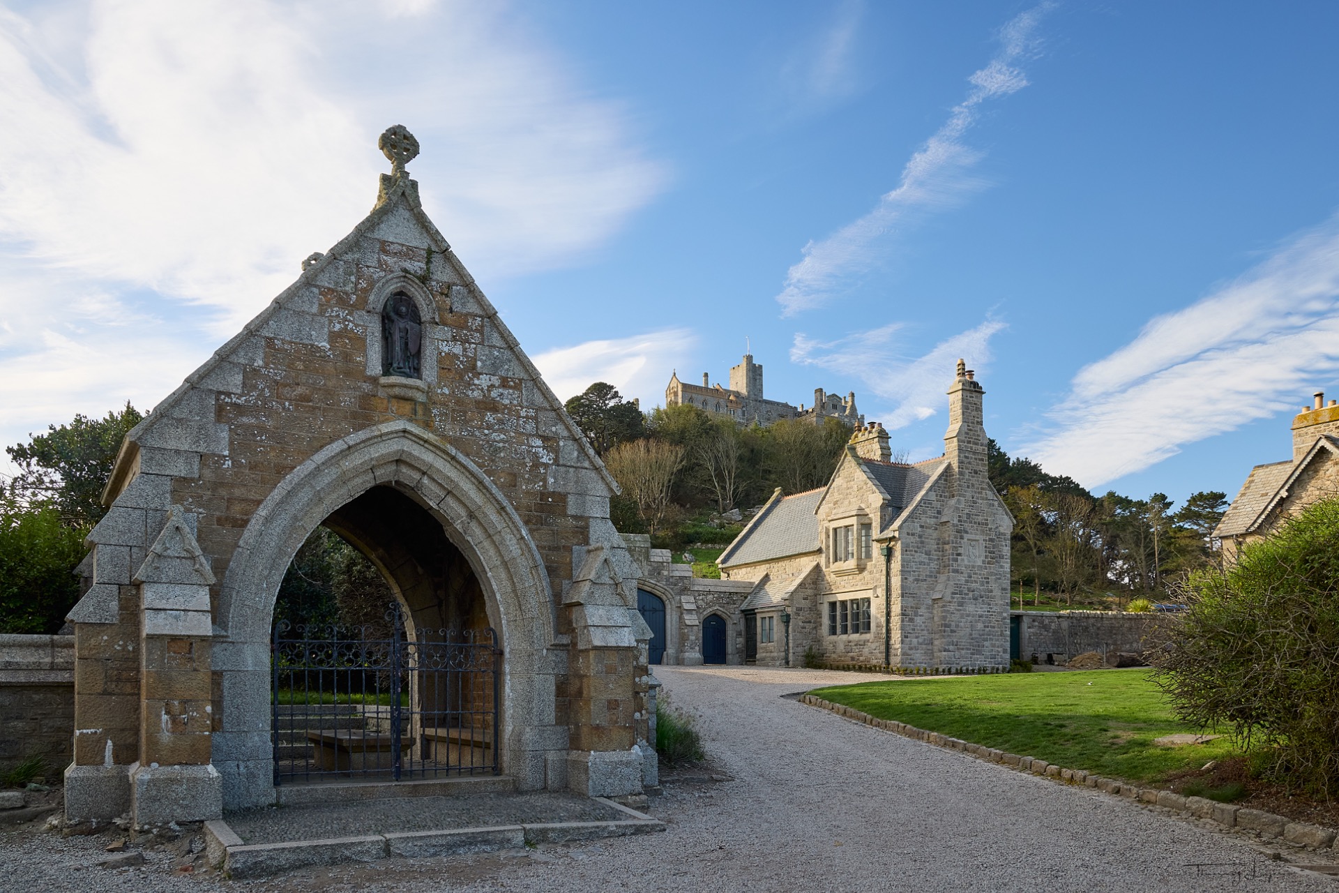 Chapelle en pierre ancienne avec portail voûté, maisons historiques au fond, ciel bleu et nuages.
