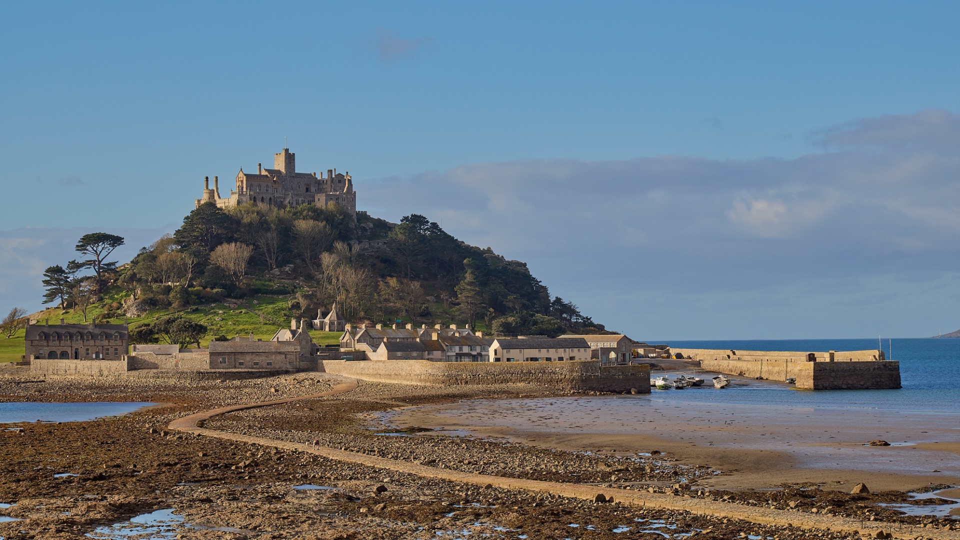 Château médiéval sur une île, maisons côtières et plage au premier plan, ciel bleu dégagé.