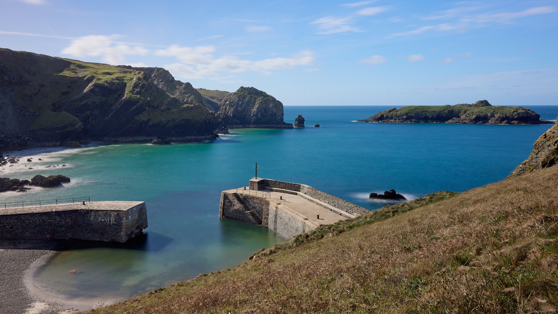 Digues d'un port dominant une crique turquoise, falaises verdoyantes et mer bleue sous un ciel clair.