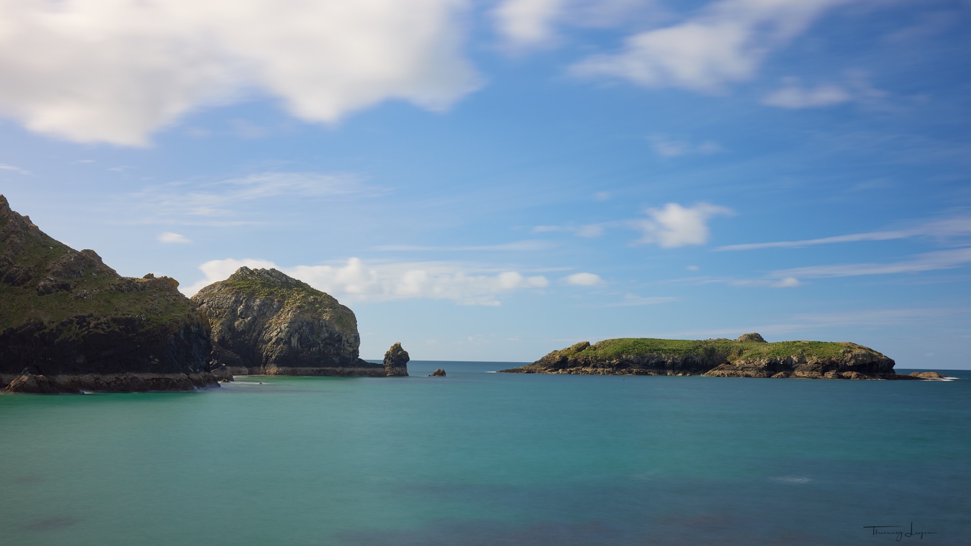 Baie aux eaux turquoises entourée de falaises rocheuses vertes, ciel bleu nuageux et rochers au loin.
