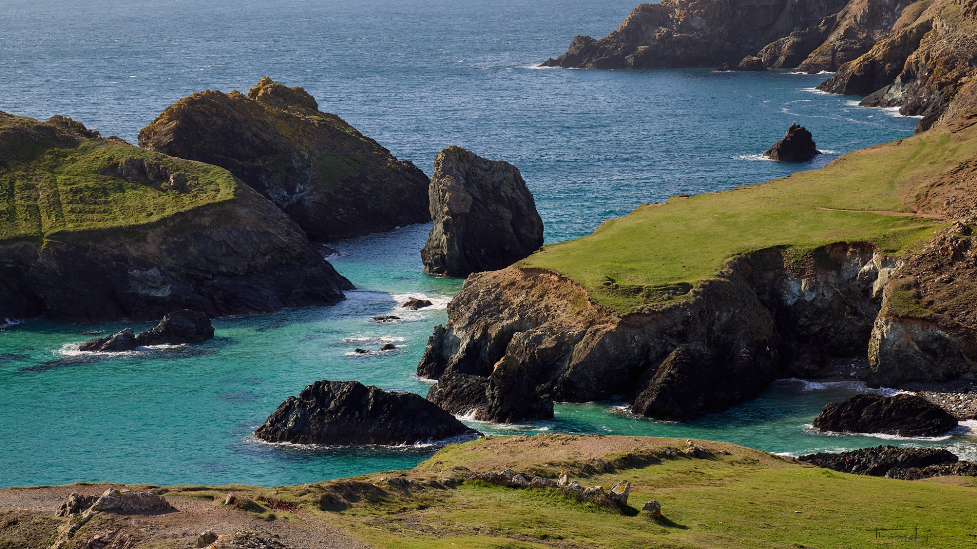 Falaises rocheuses et collines vertes surplombant une crique turquoise, vagues et rochers au large.