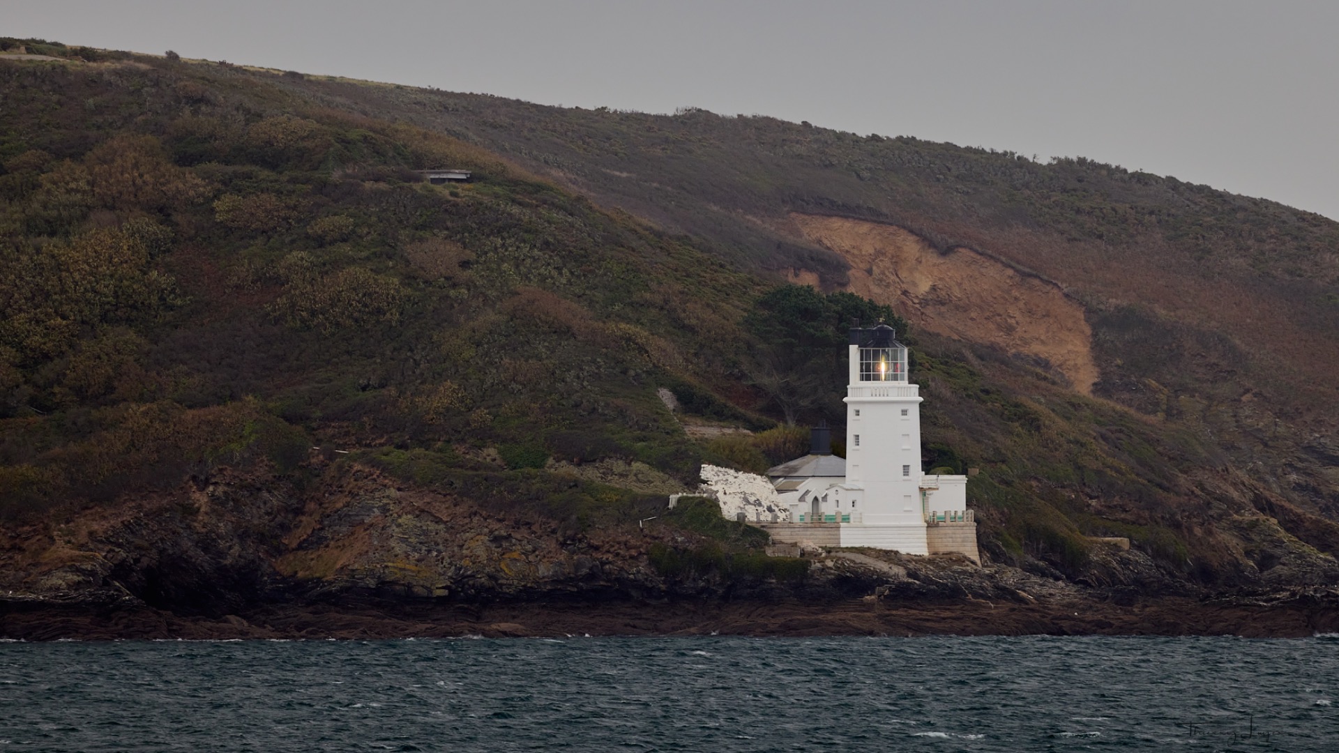 Phare blanc sur une côte rocheuse, au pied d’une colline boisée, lumière allumée au crépuscule.