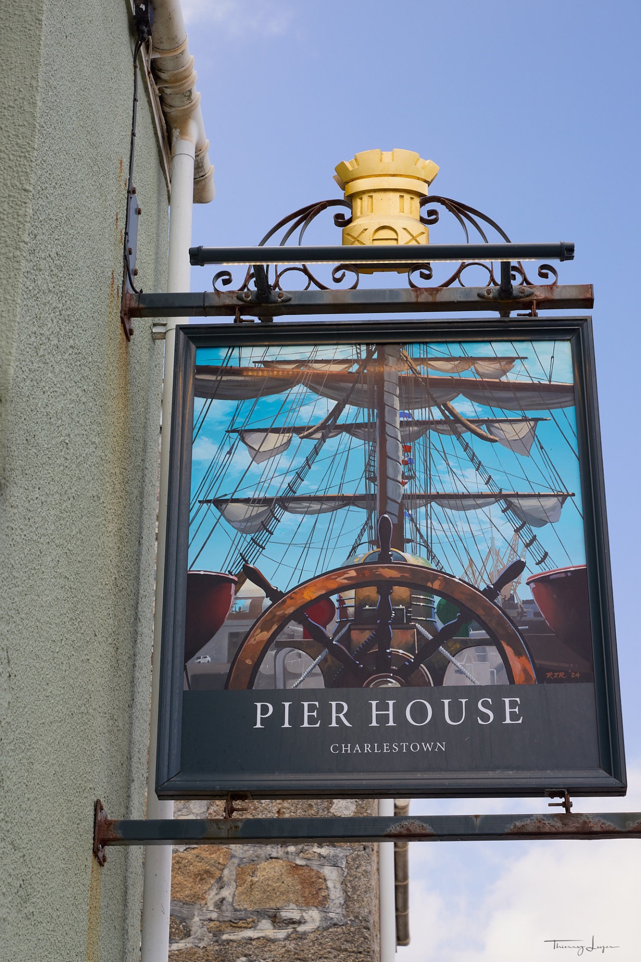 Enseigne « Pier House » avec roue et voiles miniatures, sur un mur, ciel bleu en arrière-plan.