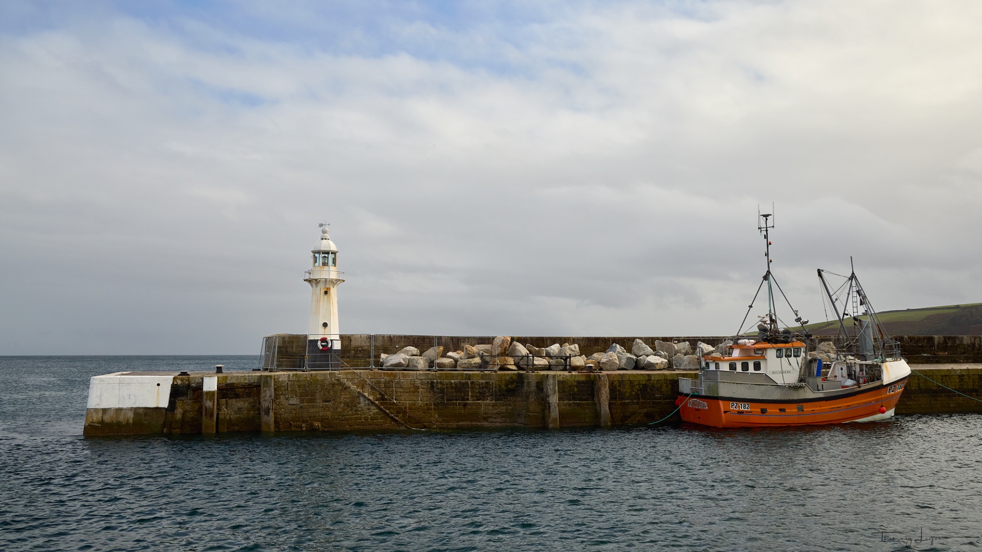 Bateau orange amarré devant un phare blanc, jetée en pierre et mer grise sous ciel nuageux.