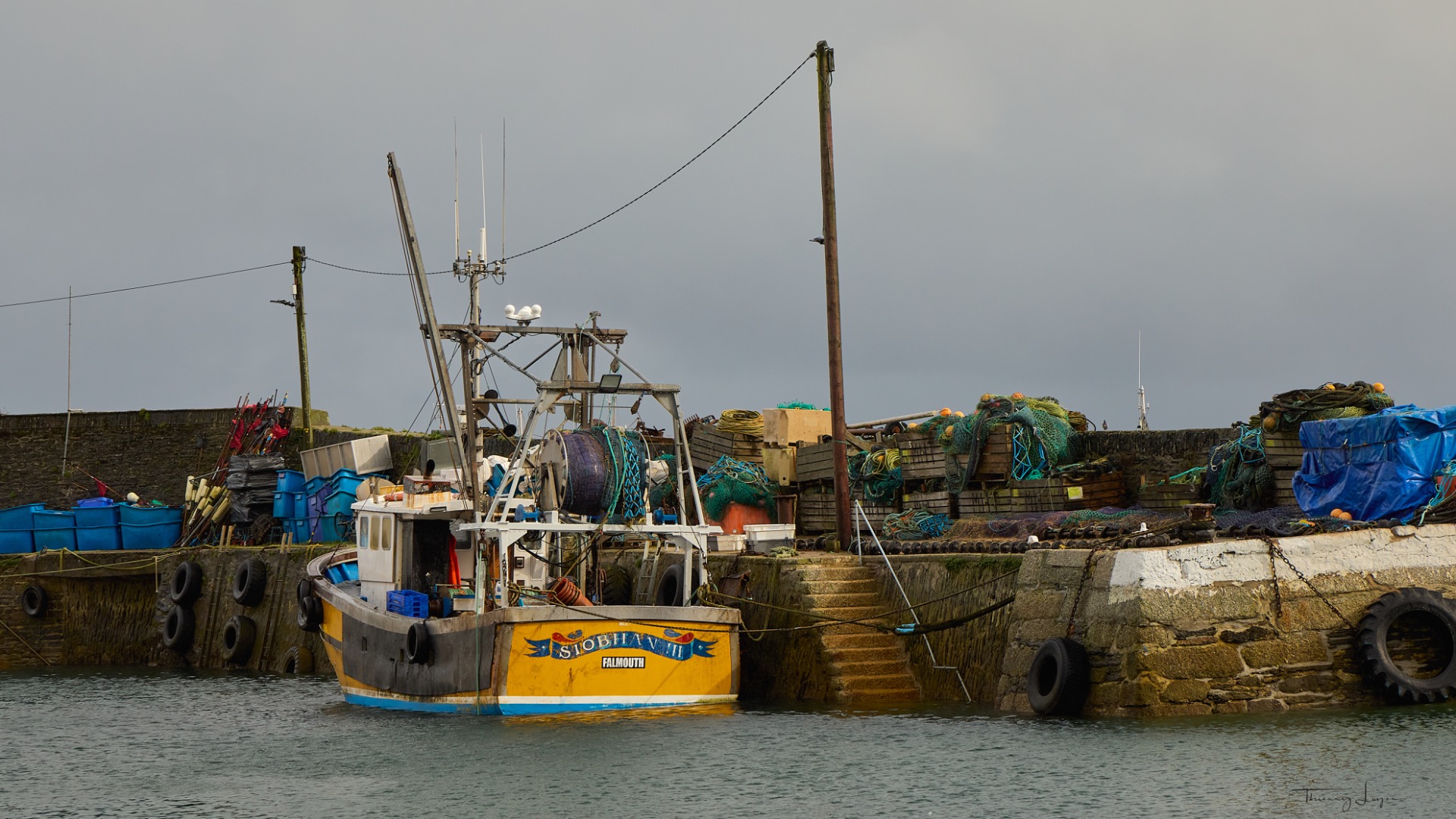 Bateau de pêche amarré au quai, filets et cordages colorés, grues et mâts contre un ciel gris.