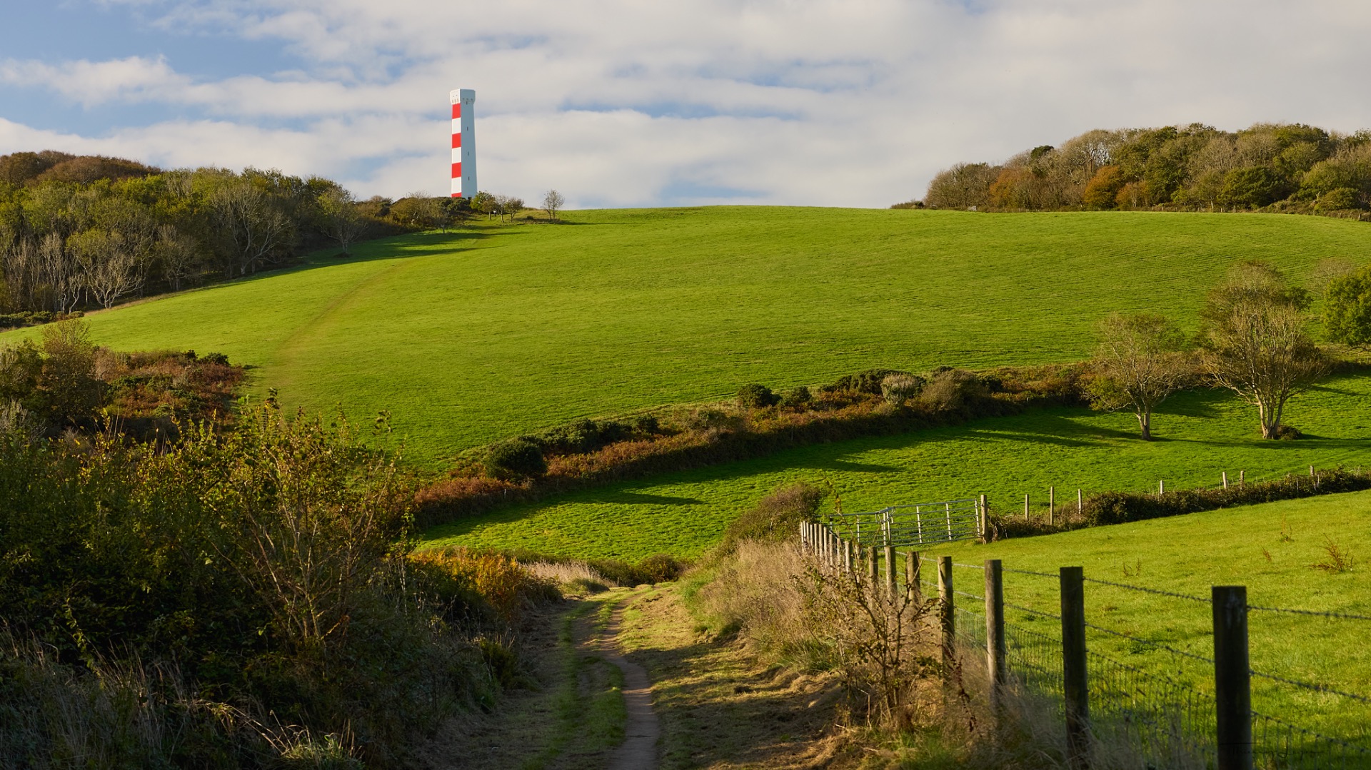 Collines verdoyantes avec une tour blanc et rouge, chemin de campagne et clôtures en bois sous ciel nuageux.