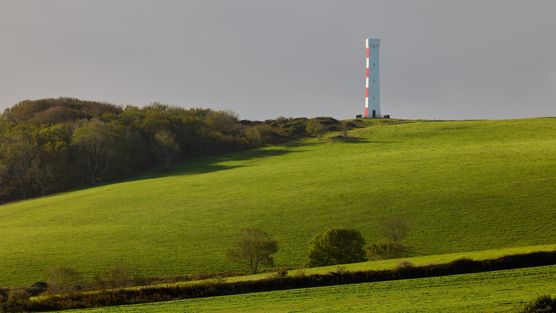 Phare blanc et rouge sur une colline, champs verts ondulés et arbres au premier plan.