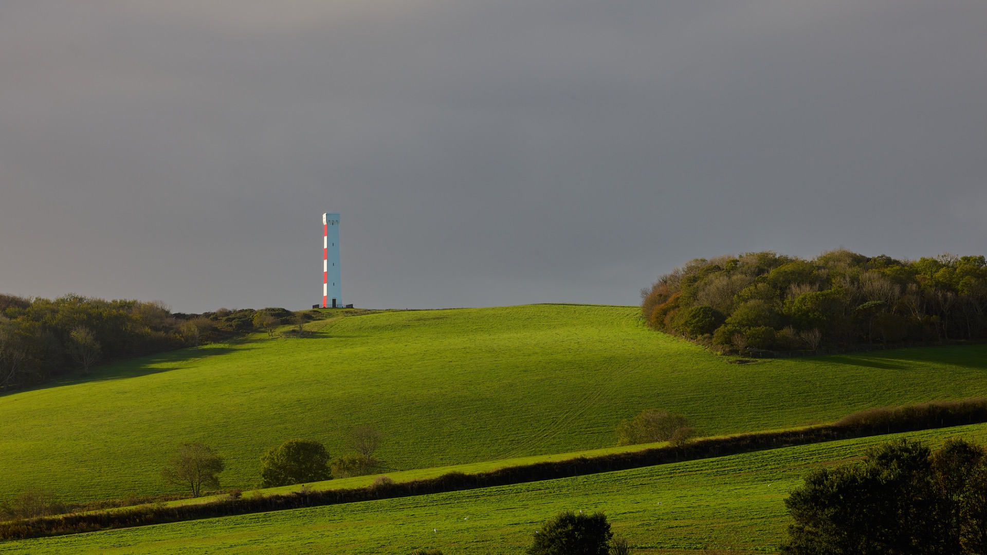 Phare blanc et rouge sur une colline, champs verts ondulés et arbres au premier plan.